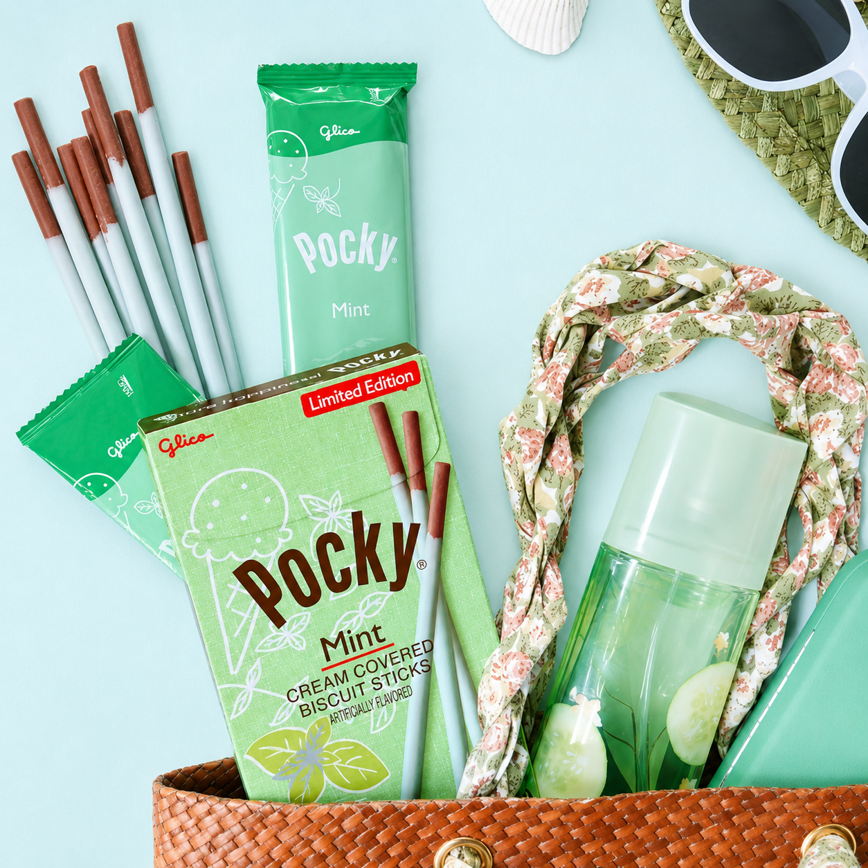 A styled flat-lay featuring a box of Pocky Mint in a woven brown tote bag, alongside a floral scarf, a clear bottle, and sunglasses.