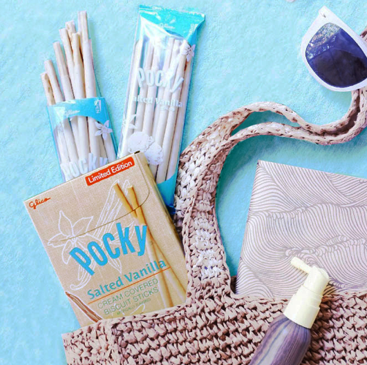 A styled flat-lay featuring a brown woven tote bag, a box of Pocky Salted Vanilla, a pair of sunglasses, and a sunblock spray bottle.
