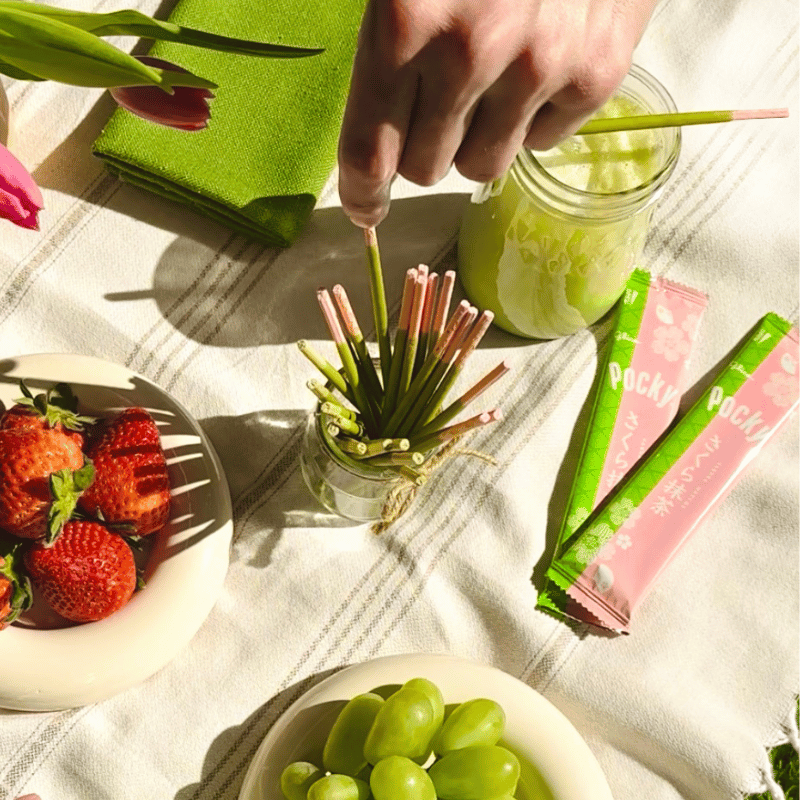 Hand reaching out to one of the Pocky Cherry Blossom Matcha sticks. A plate of strawberries, grapes, a glass of matcha latte and Pocky Cherry Blossom Matcha packets.