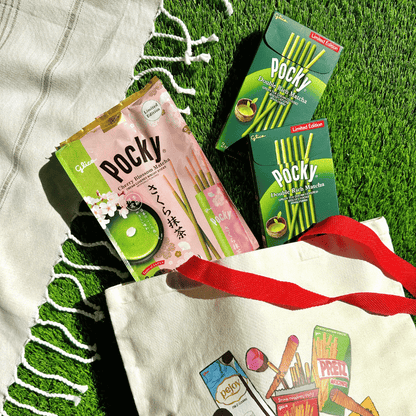 Pocky Double Rich Matcha regular-pack box and Pocky Cherry Blossom Matcha  family-pack bag coming out from a Glico tote bag on a grassy surface.