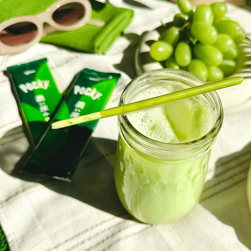 Matcha latte in a mason jar with a Pocky Double Rich Matcha stick, surrounded by grapes and Double Rich Matcha packets on a picnic blanket.