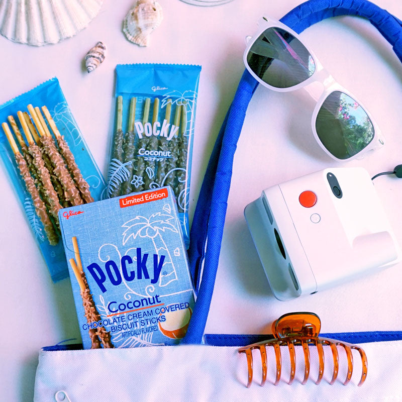 A  white tote bag filled with a package of Pocky Coconut, white sunglasses, a white instant camera, and an orange hair claw clip.