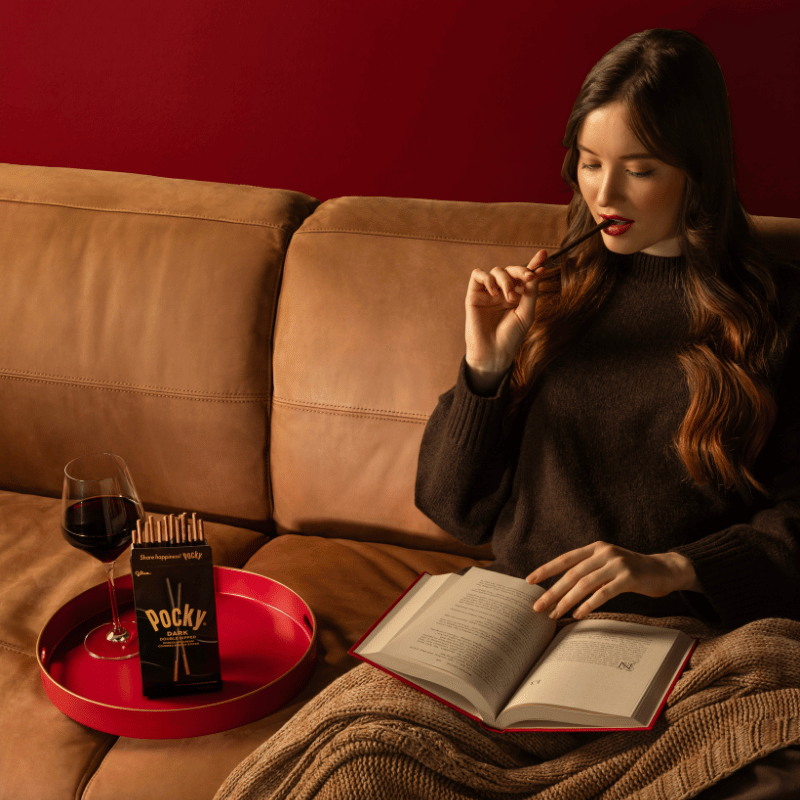 A woman sits on a couch reading a book and biting into the Pocky Dark Double Dipped stick. The next to her is a red tray holding a glass of red wine and a box of Pocky Dark Double Dipped.