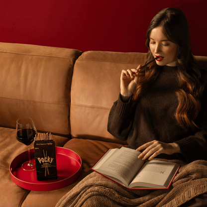 A woman sits on a couch reading a book and biting into the Pocky Dark Double Dipped stick. The next to her is a red tray holding a glass of red wine and a box of Pocky Dark Double Dipped.