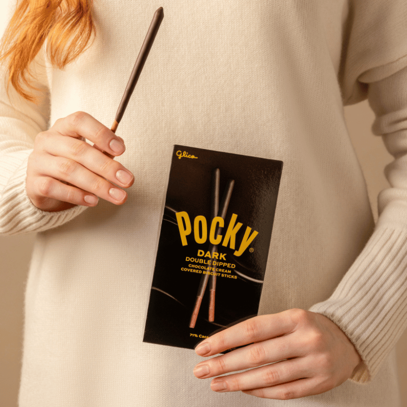 A person in a cream knit sweater holds a Pocky Dark Double Dipped stick and its black packaging box with gold lettering.